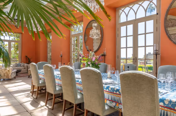 Large dining room with orange walls, tall windows, a long table covered with a patterned tablecloth, surrounded by beige chairs, and a chandelier above.