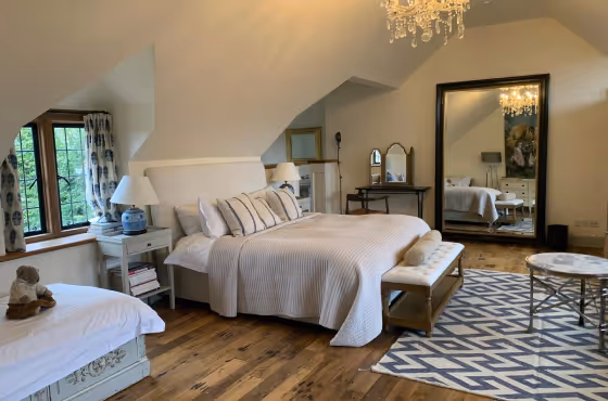 Cozy bedroom with a large bed, white bedding, wooden floor, a geometric patterned rug, chaise lounge, and a tall mirror reflecting a chandelier.