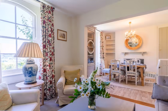Bright living and dining area with floral curtains, armchairs, a vase of daisies on a coffee table, and a wooden dining table with chairs under a chandelier.