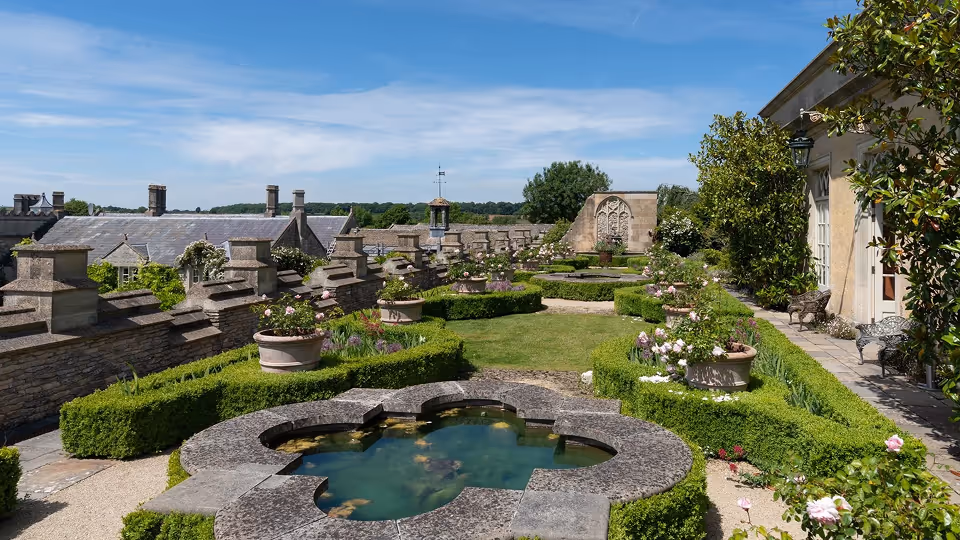 Formal garden with a cloverleaf-shaped stone pond in the foreground, manicured hedges, potted plants with flowers, and historic stone buildings under a blue sky.