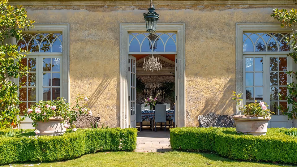 Elegant garden patio with trimmed hedges, potted flowers, and an open door leading to a dining room with a chandelier.