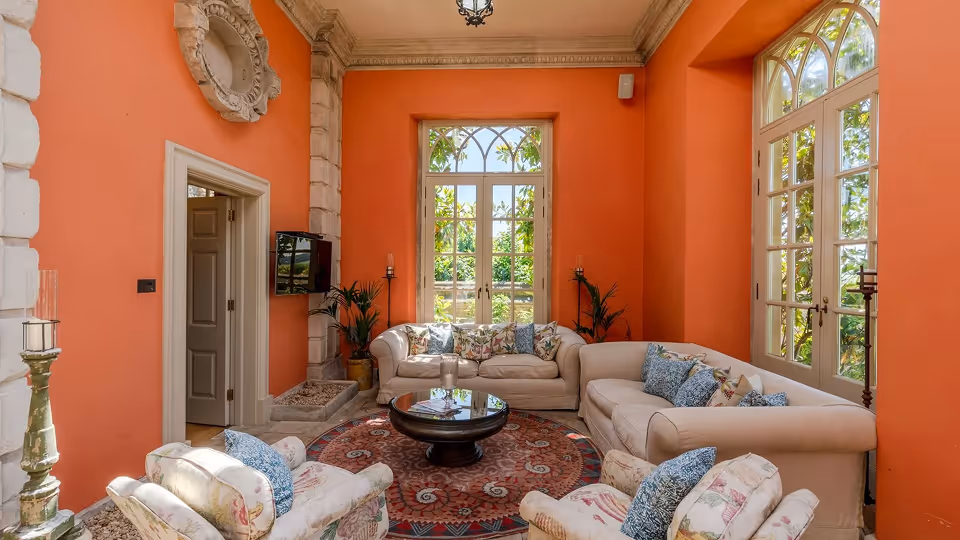 Cozy living room with vibrant orange walls, large arched windows, floral and patterned sofas around a circular wooden coffee table on a red ornate rug.
