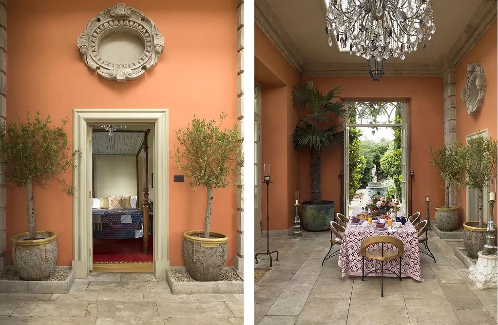 Elegant outdoor dining area with a table set for breakfast under a crystal chandelier, flanked by large potted plants, and a doorway leading to a bedroom with a four-poster bed.