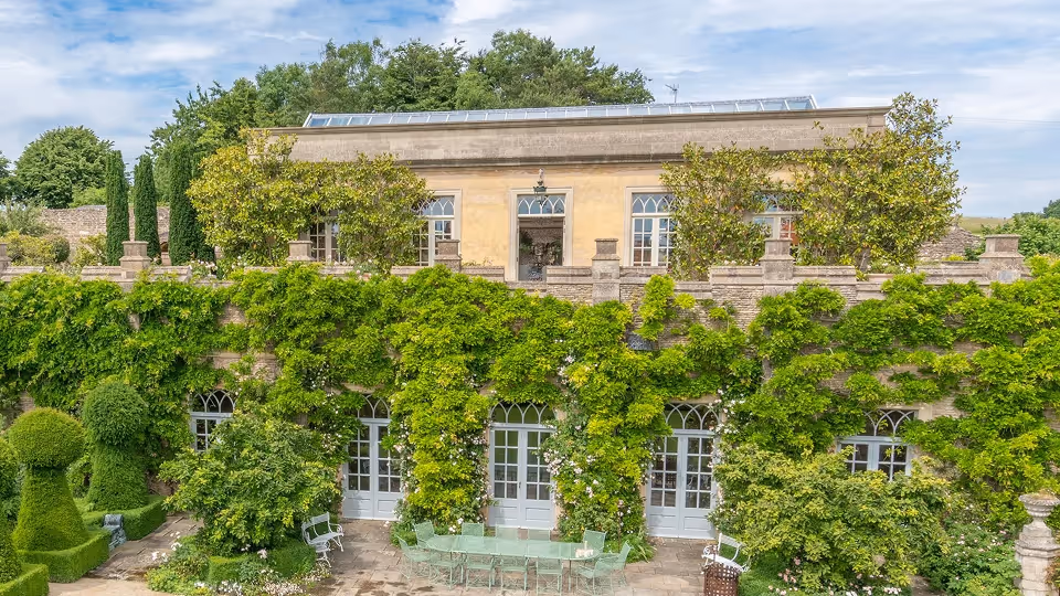 Historic stone building with upper terrace, covered in green ivy and surrounded by manicured garden plants.