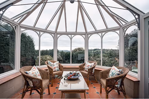 Bright sunroom with glass ceiling and walls, furnished with four wicker chairs with floral cushions around a tiled coffee table with a fruit bowl.