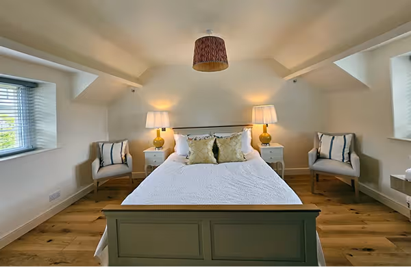 Bright bedroom with a white bed centered between two nightstands with lamps and two armchairs by windows on either side.