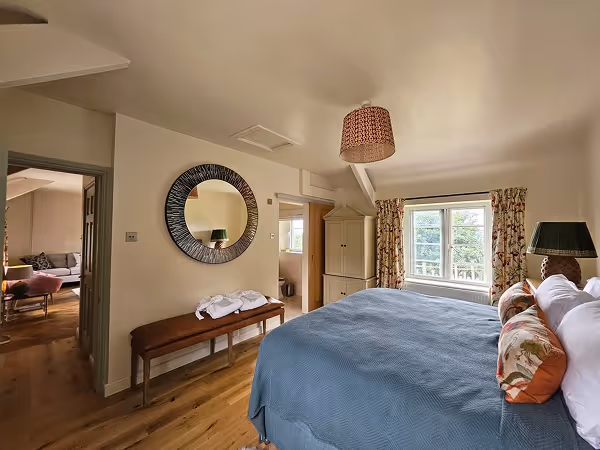 Cozy bedroom with a blue bedspread, floral pillows, a round wall mirror, and a window with patterned curtains.