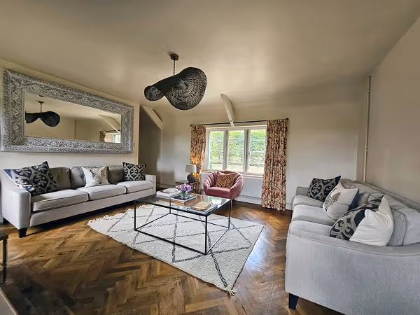 Living room with two gray sofas, a pink armchair, a glass coffee table on a geometric patterned rug, wooden parquet floor, large window with floral curtains, and a decorative wall mirror.