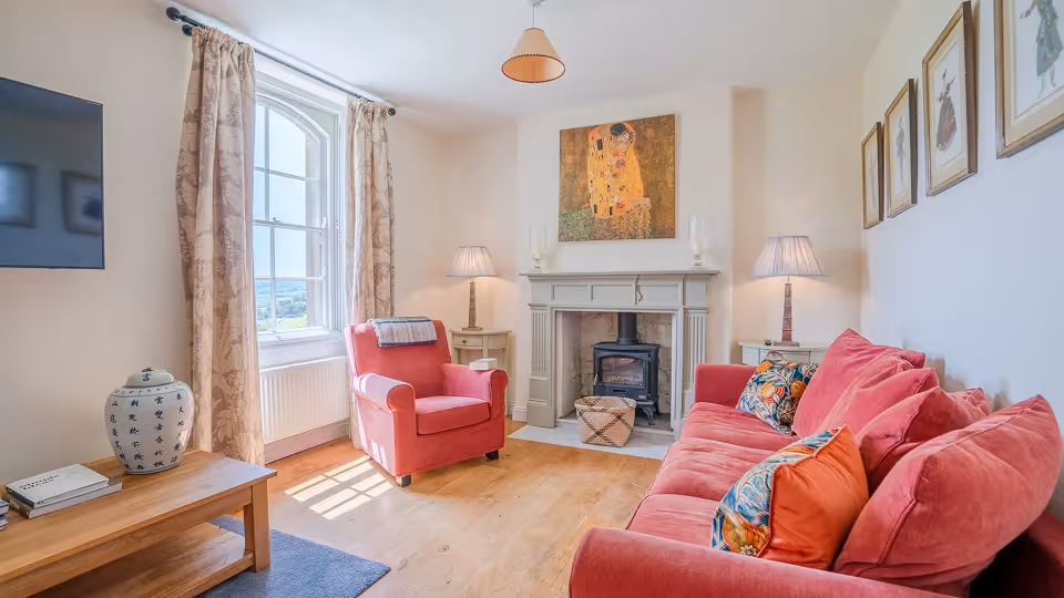Cozy living room with a red armchair and sofa, wooden floor, two side tables with lamps, fireplace, and framed artwork on the walls.