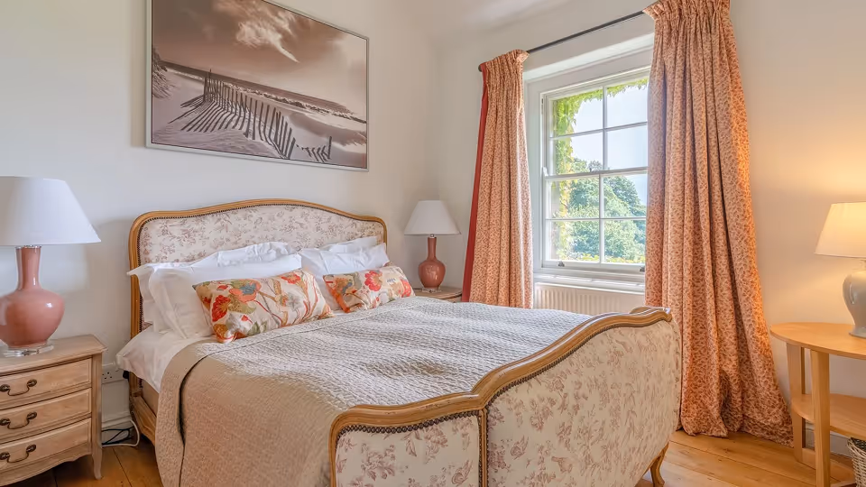 Cozy bedroom with a floral upholstered bed, patterned curtains, wooden side tables, and lamps under a window.