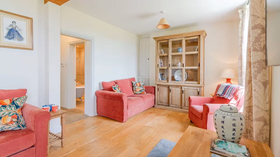 Cozy living room with pink armchairs and sofa, wooden floor, glass-front cabinet, and decorative cushions.