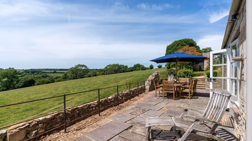 Stone patio with wooden chairs, a table under a blue umbrella, and rolling green fields under a clear sky.