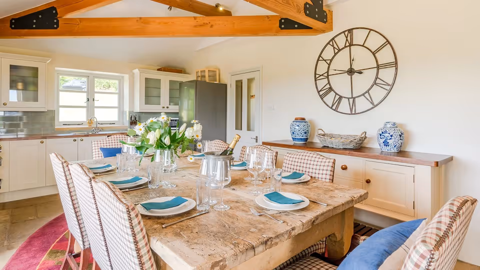 Rustic dining table set with white plates, blue napkins, glassware, and a flower centerpiece in a bright kitchen with wooden beams and a large wall clock.
