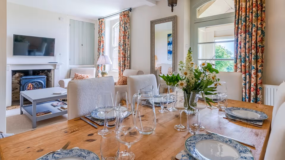 Cozy dining area with a wooden table set for six, floral centerpiece, white cushioned chairs, and a living room with a fireplace and television.