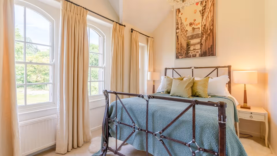 Bright bedroom with a metal bedframe, green bedspread, multiple pillows, beige curtains, and two bedside lamps.