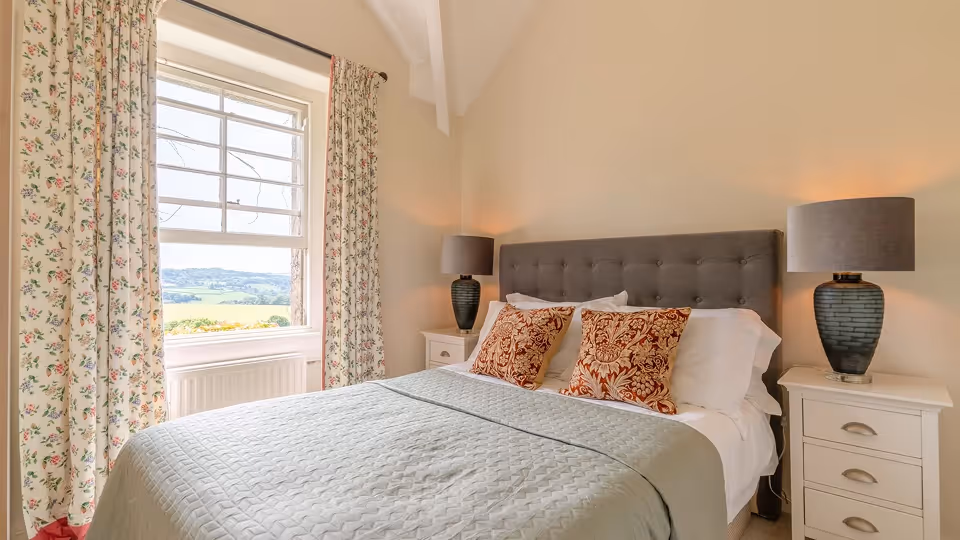 Cozy bedroom with a large bed, patterned pillows, two bedside tables with matching lamps, and floral curtains framing a window with a countryside view.