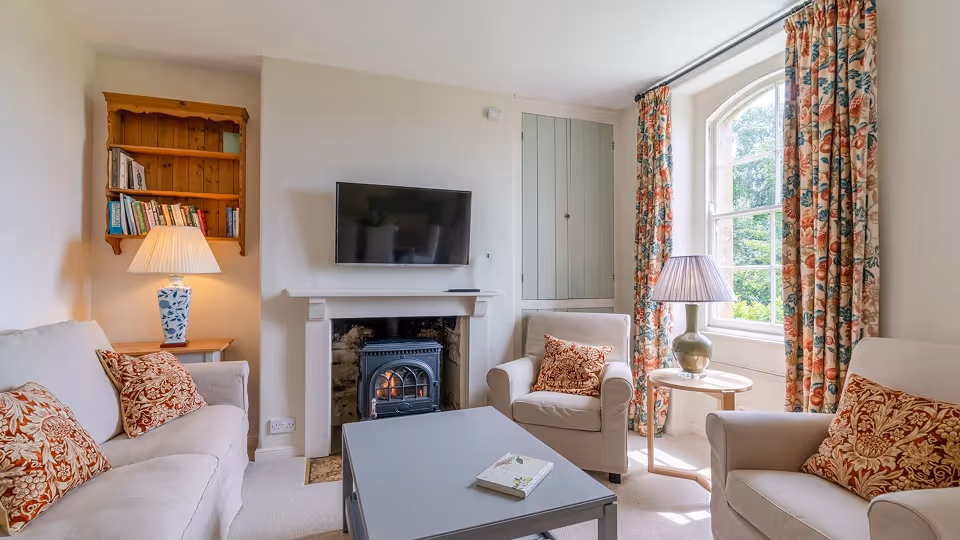 Cozy living room with beige sofas, patterned cushions, floral curtains, a gray coffee table, a lit fireplace under a mounted TV, and table lamps.