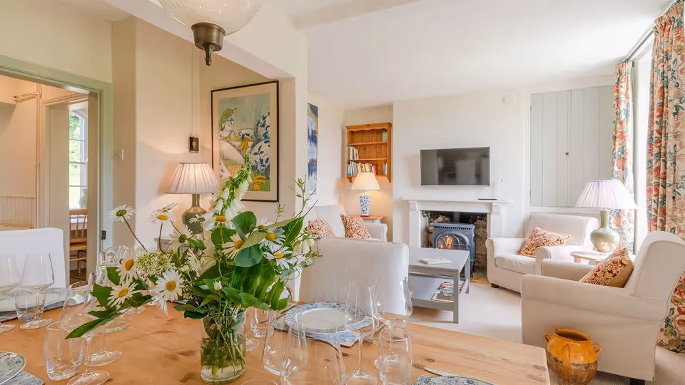 Cozy living and dining area with white armchairs, floral curtains, a wooden table set with plates and glasses, and a vase of daisies in the foreground.
