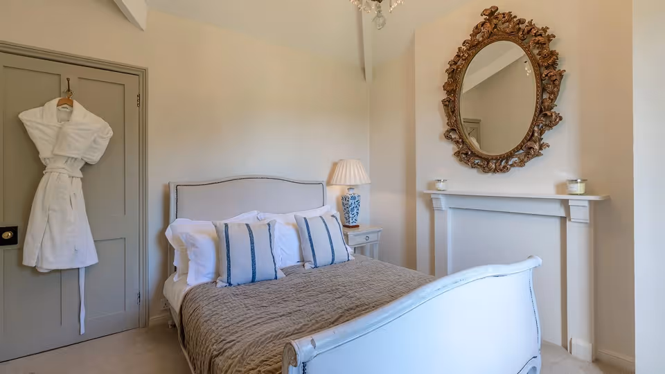 Cozy bedroom with a white and blue bed, decorative pillows, a bedside lamp, a white robe hanging on a gray door, and an ornate oval mirror above a faux fireplace.