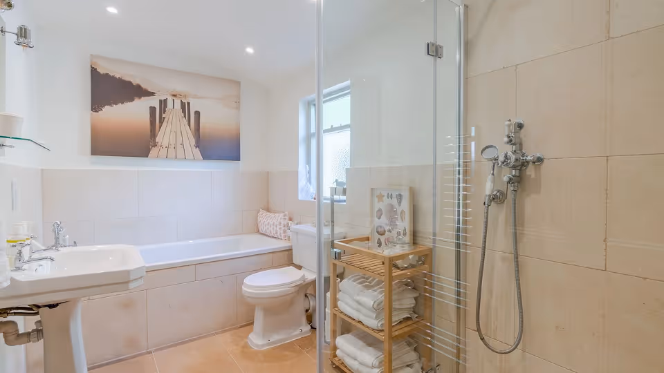 Bright bathroom with a white bathtub, toilet, pedestal sink, glass shower enclosure, and wooden shelf with towels.