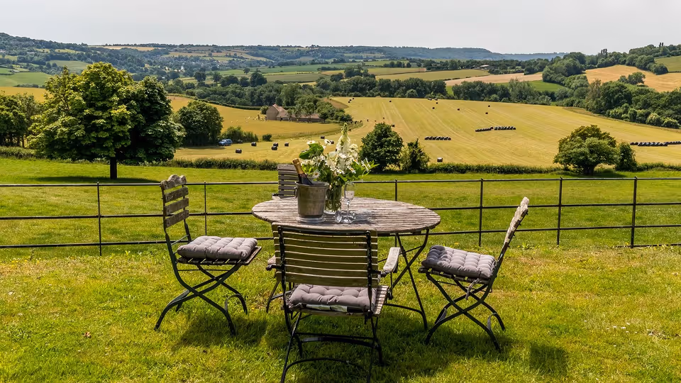 Round wooden table with four cushioned chairs on green grass overlooking fields and rolling hills under a cloudy sky.