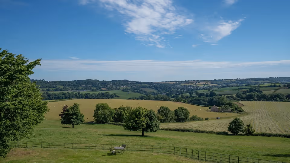 Expansive green countryside with scattered trees, fenced grassy fields, and rolling hills under a blue sky with clouds.