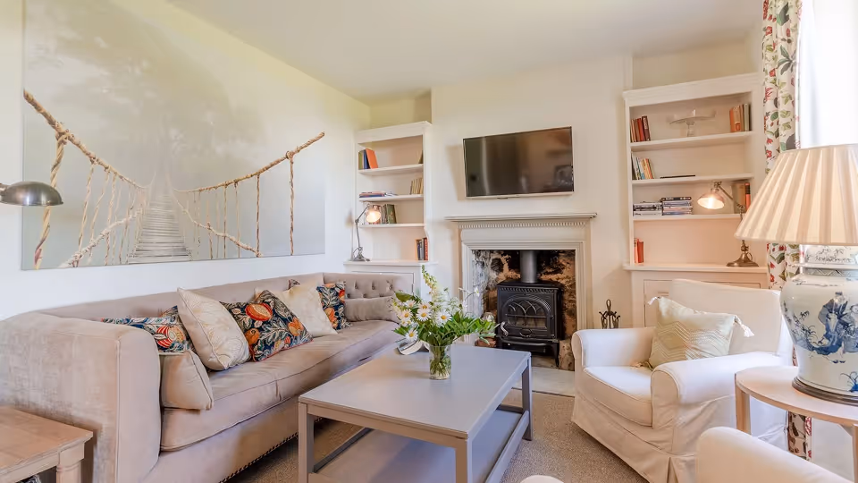Cozy living room with beige sofa, patterned cushions, armchair, coffee table with flowers, fireplace, TV, and bookshelves.