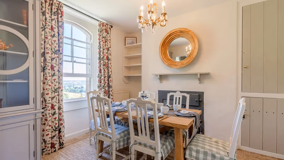 Bright dining room with wooden table set for four, white chairs with checkered cushions, floral curtains, and a round wooden mirror above a small fireplace.