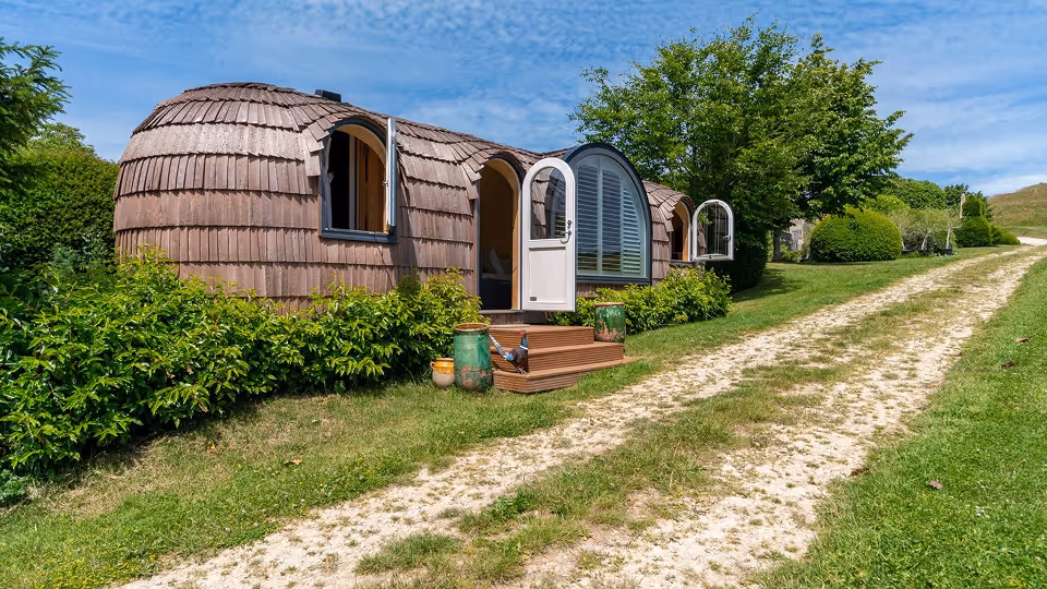 Unique small curved wooden house with open doors and windows along a grassy dirt path in a lush green landscape under a blue sky.