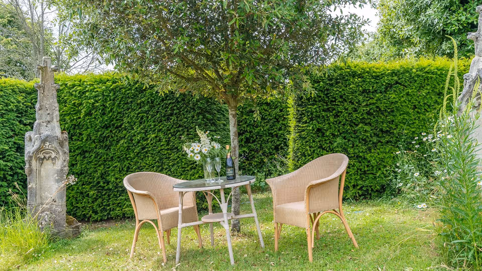 Outdoor garden scene with two wicker chairs and a small round table under a tree, decorated with flowers and a bottle on the table.
