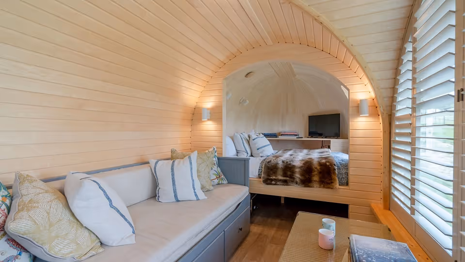 Cozy small living space with wooden walls, a beige sofa with cushions, a coffee table, and a bedroom area visible through an arched opening.