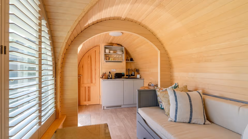 Cozy wooden interior of a small living space featuring a cushioned bench with pillows, a kitchenette, and a window with white blinds.