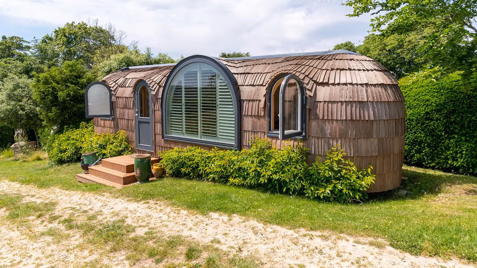 Small curved wooden cabin with rounded windows and shingle siding surrounded by greenery and grass.