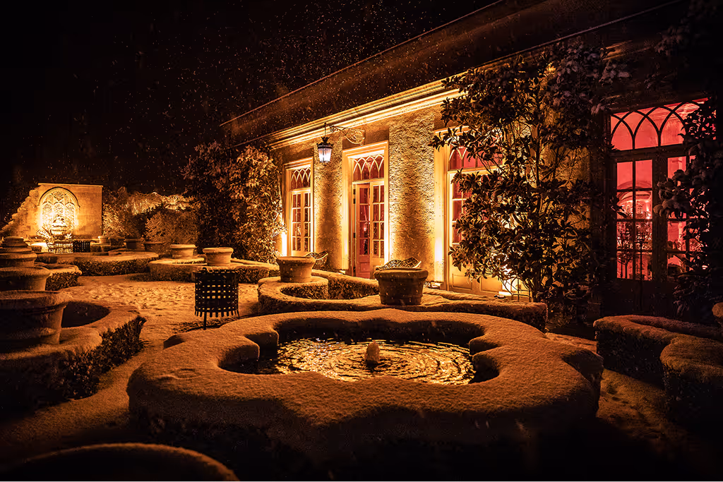 Snow-covered courtyard at night with illuminated building and water fountain surrounded by bushes and large planters.