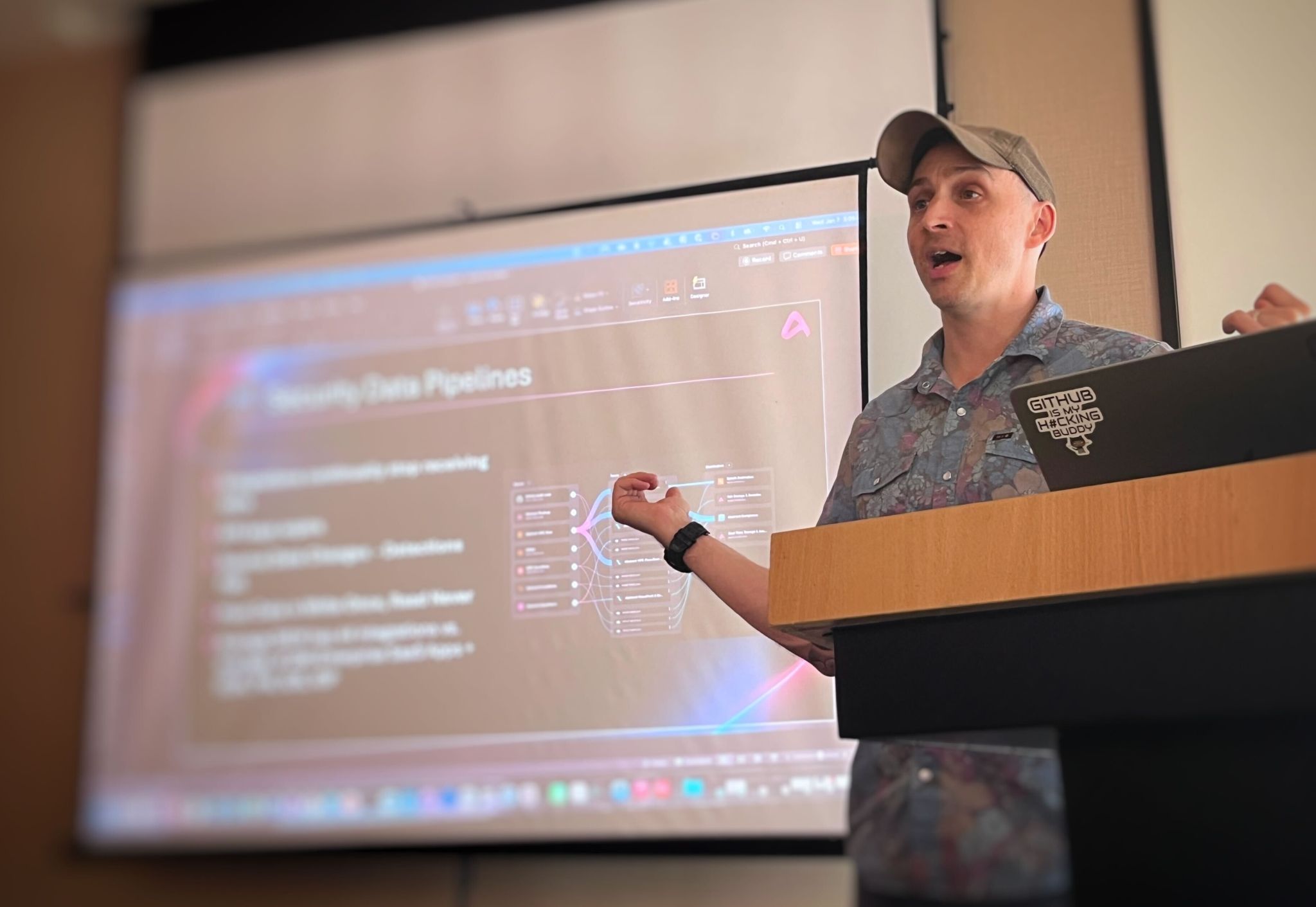 Man wearing a cap and floral shirt giving a presentation on securing data pipelines, standing behind a podium with a laptop sticker that reads 'GITHUB IS MY HACKING BUDDY'.