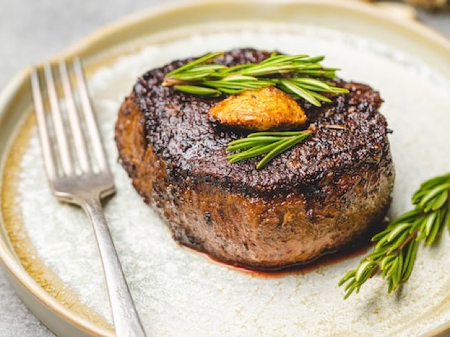 A thick, seared filet mignon steak resting on a light-colored plate. The steak has a dark, caramelized crust and is topped with a pat of melting herb butter and fresh rosemary sprigs. A silver fork sits to the left, and additional rosemary is visible in the background.