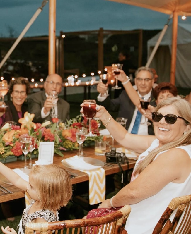 Group of adults and a child seated at a long outdoor table, smiling and raising glasses in a toast during an evening celebration under a tent with string lights.