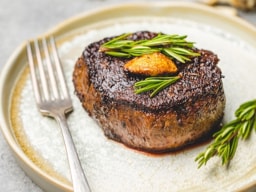 A thick, seared filet mignon steak resting on a light-colored plate. The steak has a dark, caramelized crust and is topped with a pat of melting herb butter and fresh rosemary sprigs. A silver fork sits to the left, and additional rosemary is visible in the background.
