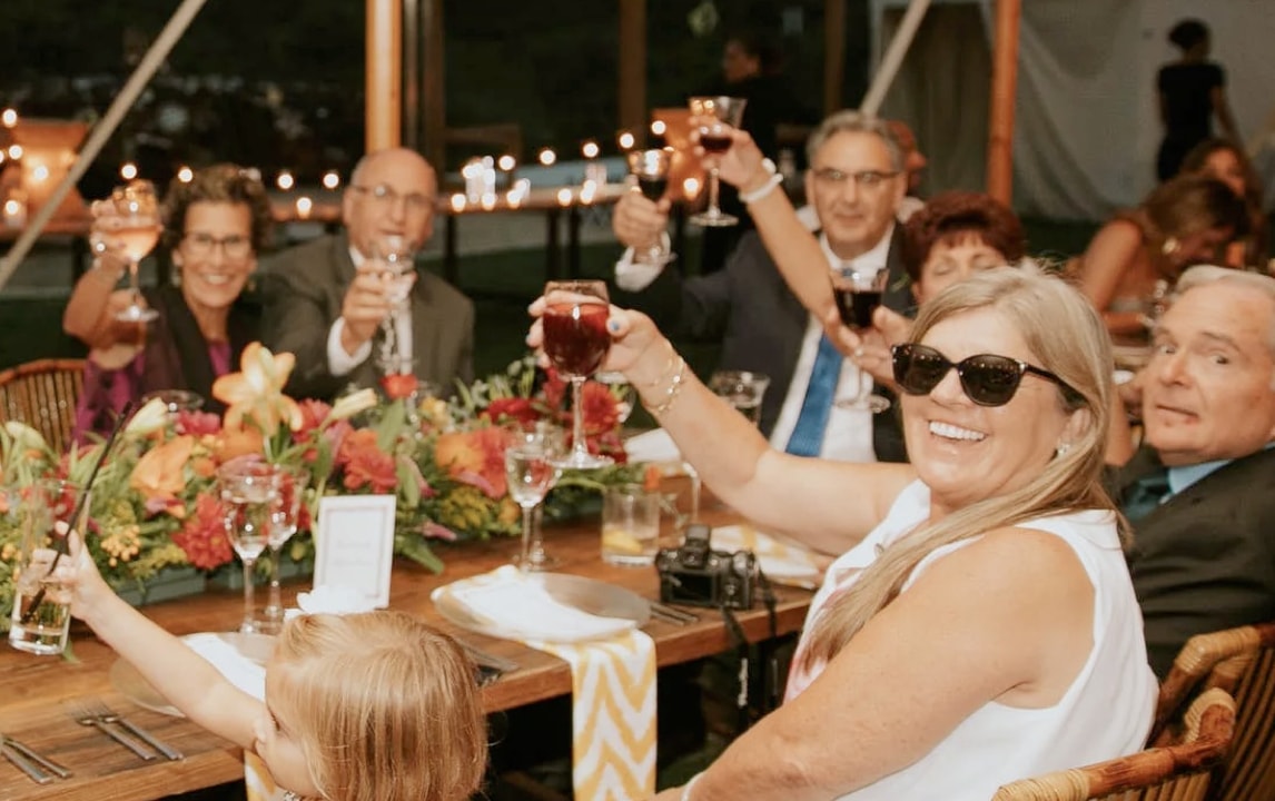 Group of adults and a child seated at a long outdoor table, smiling and raising glasses in a toast during an evening celebration under a tent with string lights.
