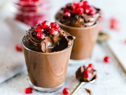 Two small glass cups filled with smooth, dark chocolate mousse. Each serving is topped with a generous cluster of bright red pomegranate seeds. A gold spoon with a small bite of mousse sits in the foreground on a light, textured surface.