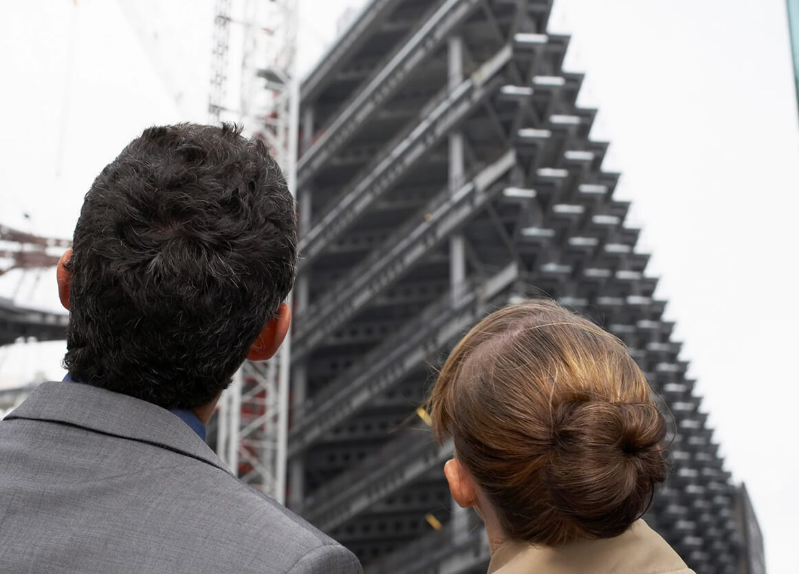 Two people in business attire look up at a tall, unfinished building under construction.