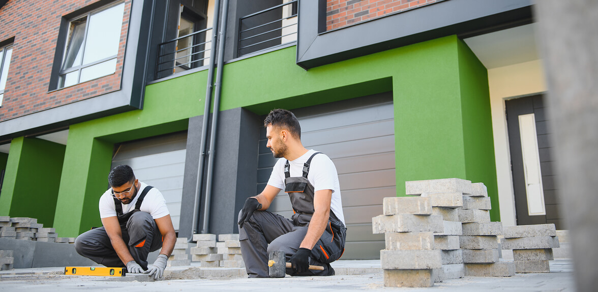 Two construction workers in overalls are laying pavement stones in front of a modern building with green accents.