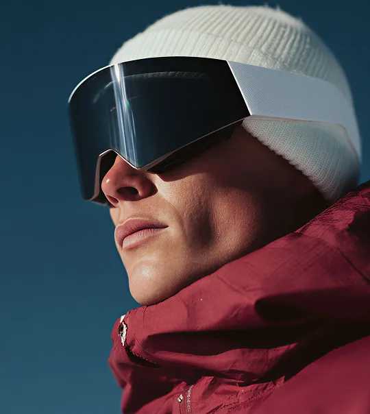 Close-up of a person wearing a white knit beanie, large futuristic black visor sunglasses, and a red jacket against a clear blue sky.