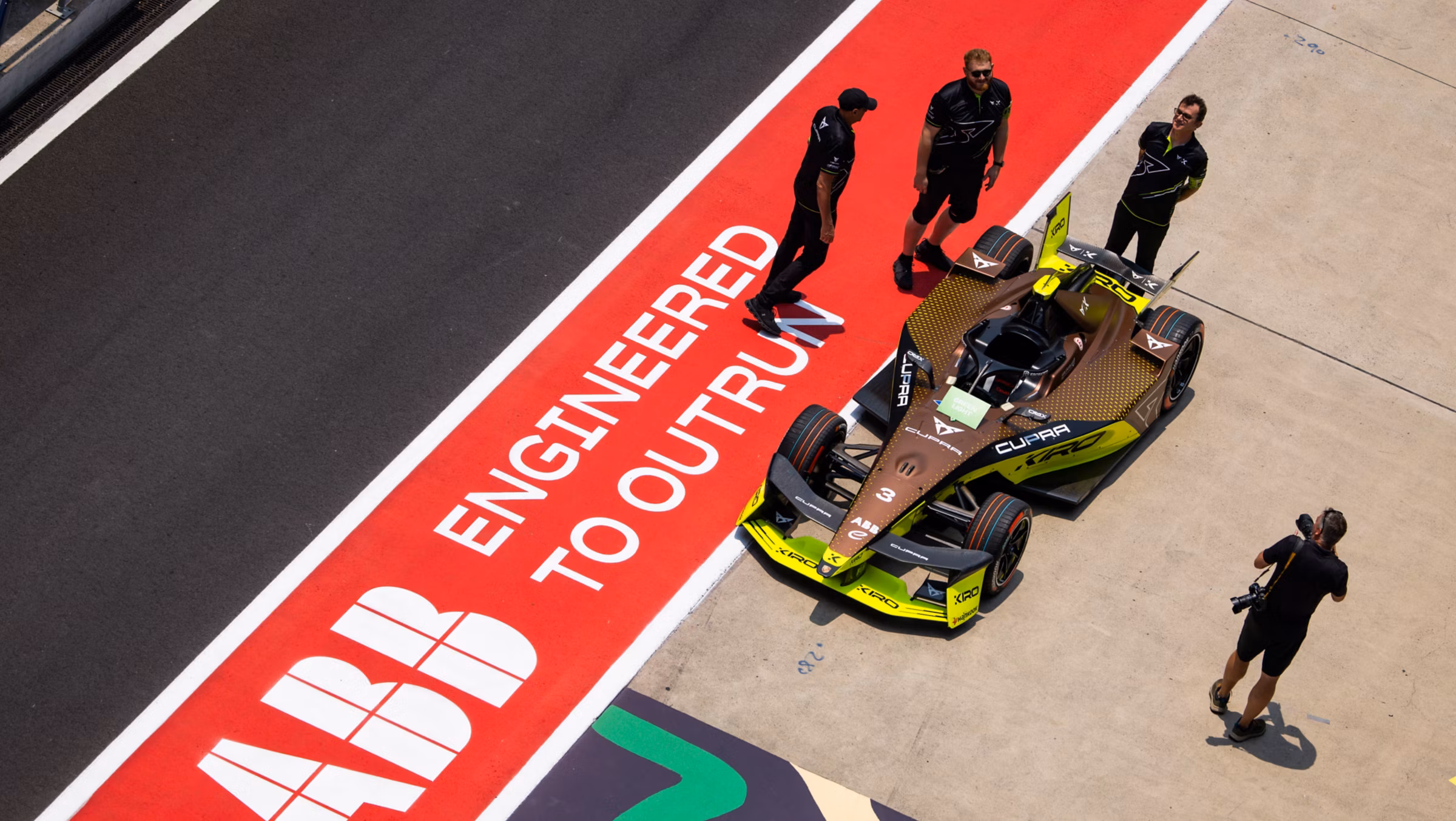 Overhead view of a brown and neon-yellow Cupra Formula E race car on a pit lane with four team members and one photographer nearby.