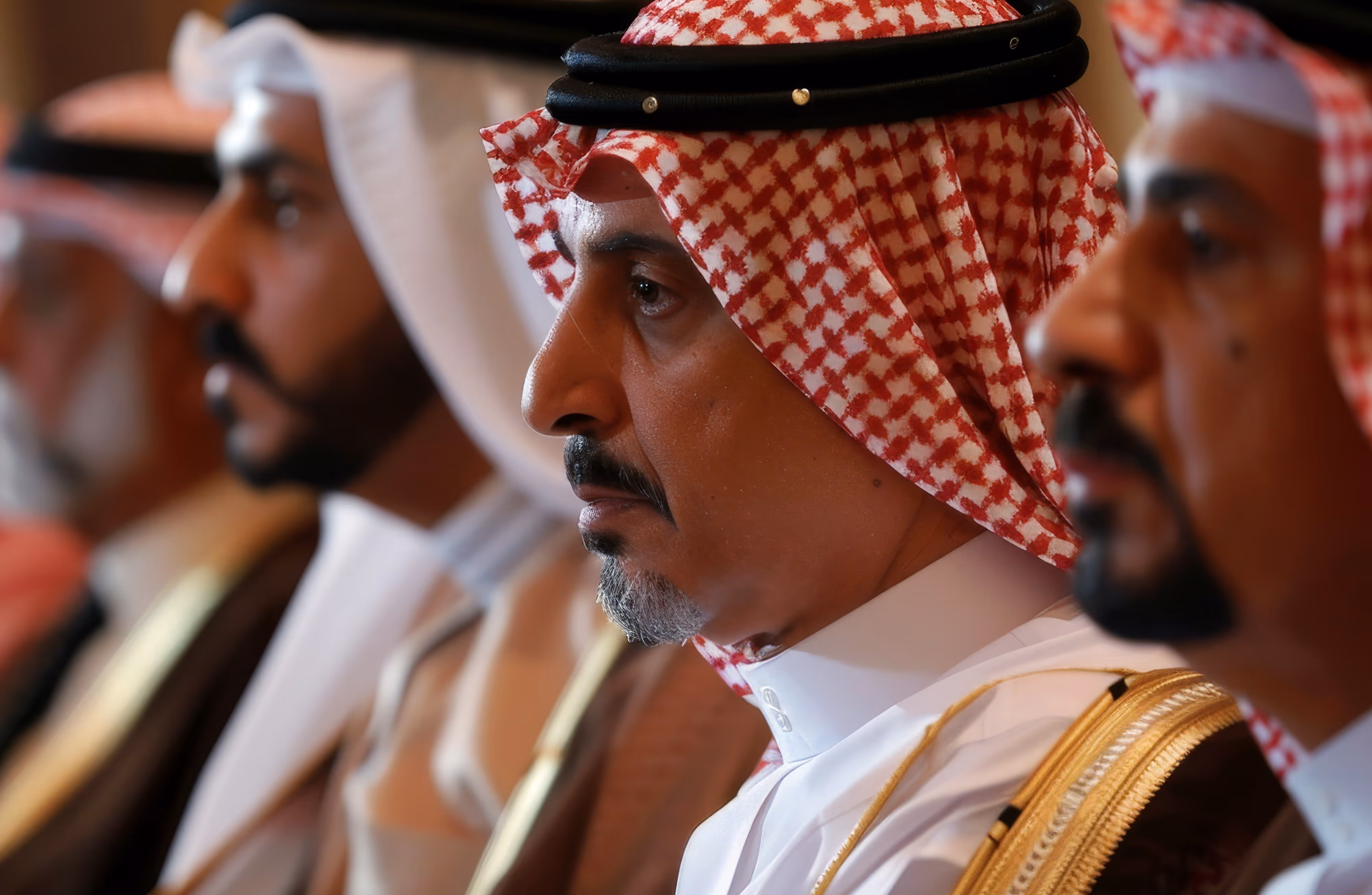 Four Middle Eastern men in traditional attire sitting in a row, with the foremost man wearing a red and white checkered headscarf.