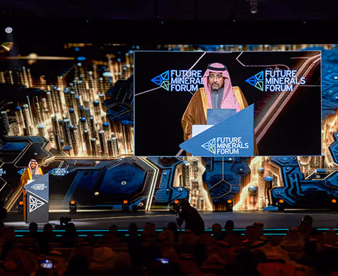 Speaker in traditional Middle Eastern attire delivering a speech at the Future Minerals Forum with a large screen backdrop displaying the event logo.