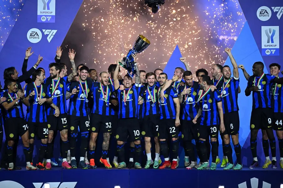 Football team wearing black and blue striped jerseys celebrating with a trophy on a stage with confetti and fireworks in the background.