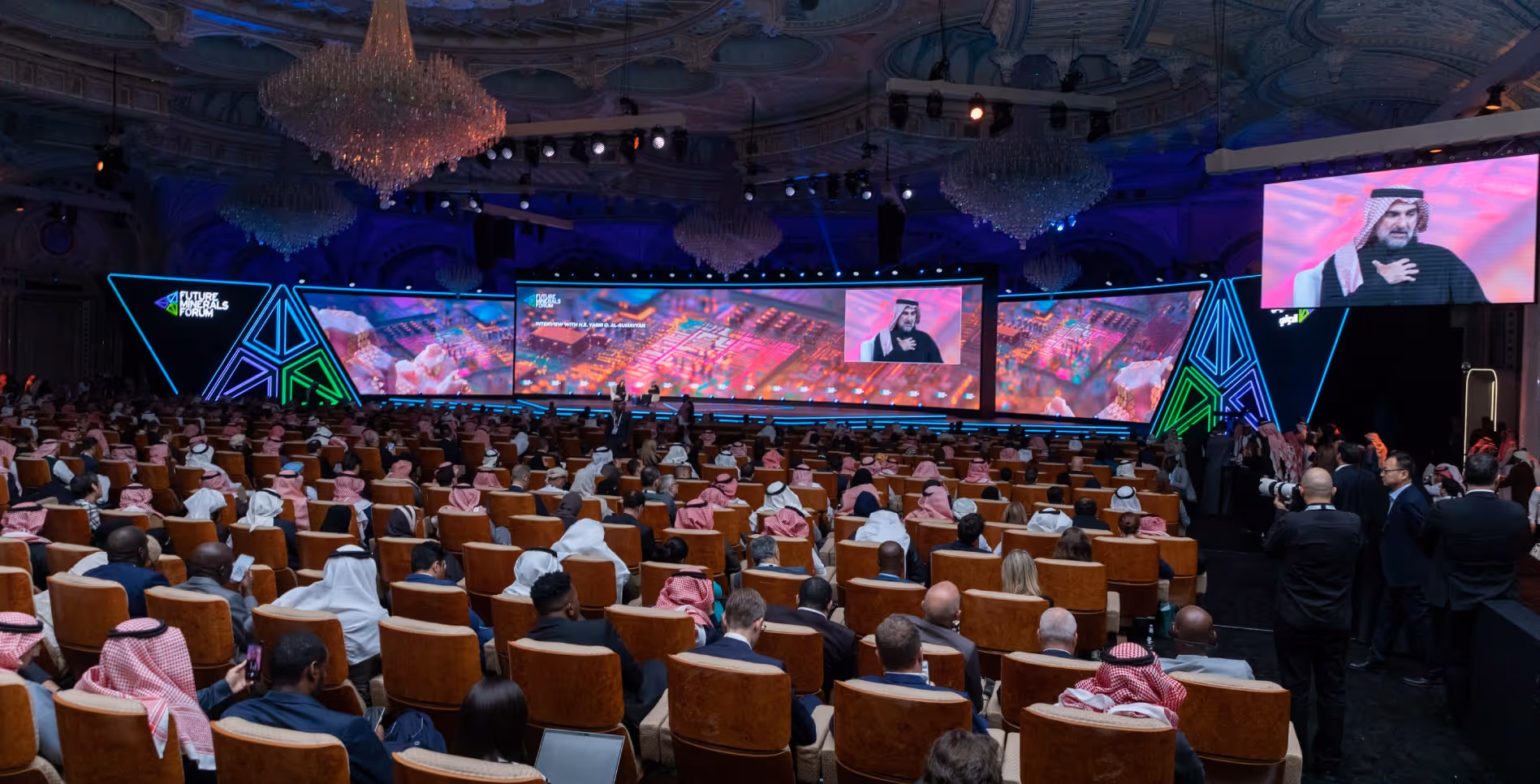 Large conference hall filled with attendees watching a speaker on a wide stage with colorful digital displays and chandeliers above.