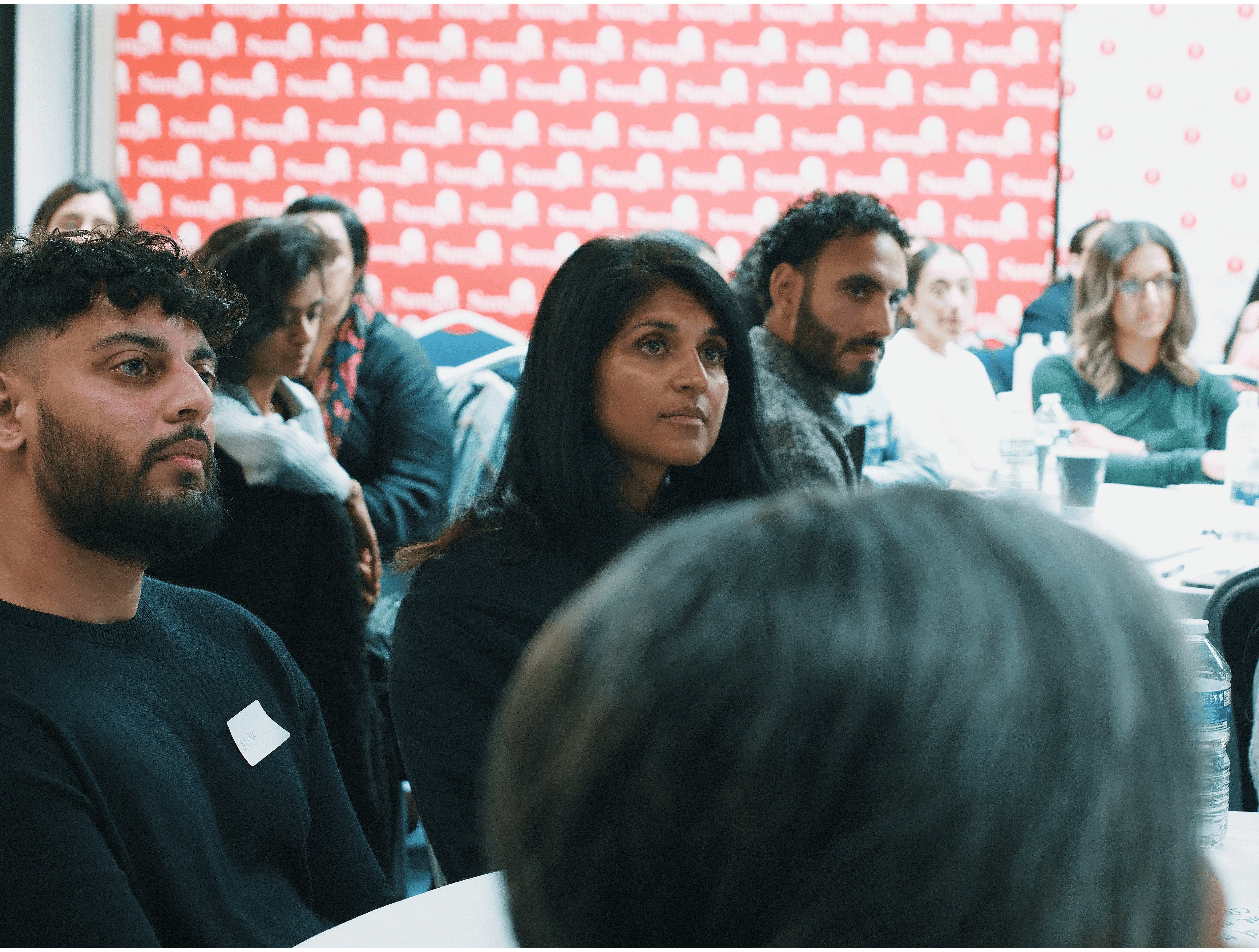 A diverse group of people attentively listening at an indoor event with a red and white patterned backdrop.