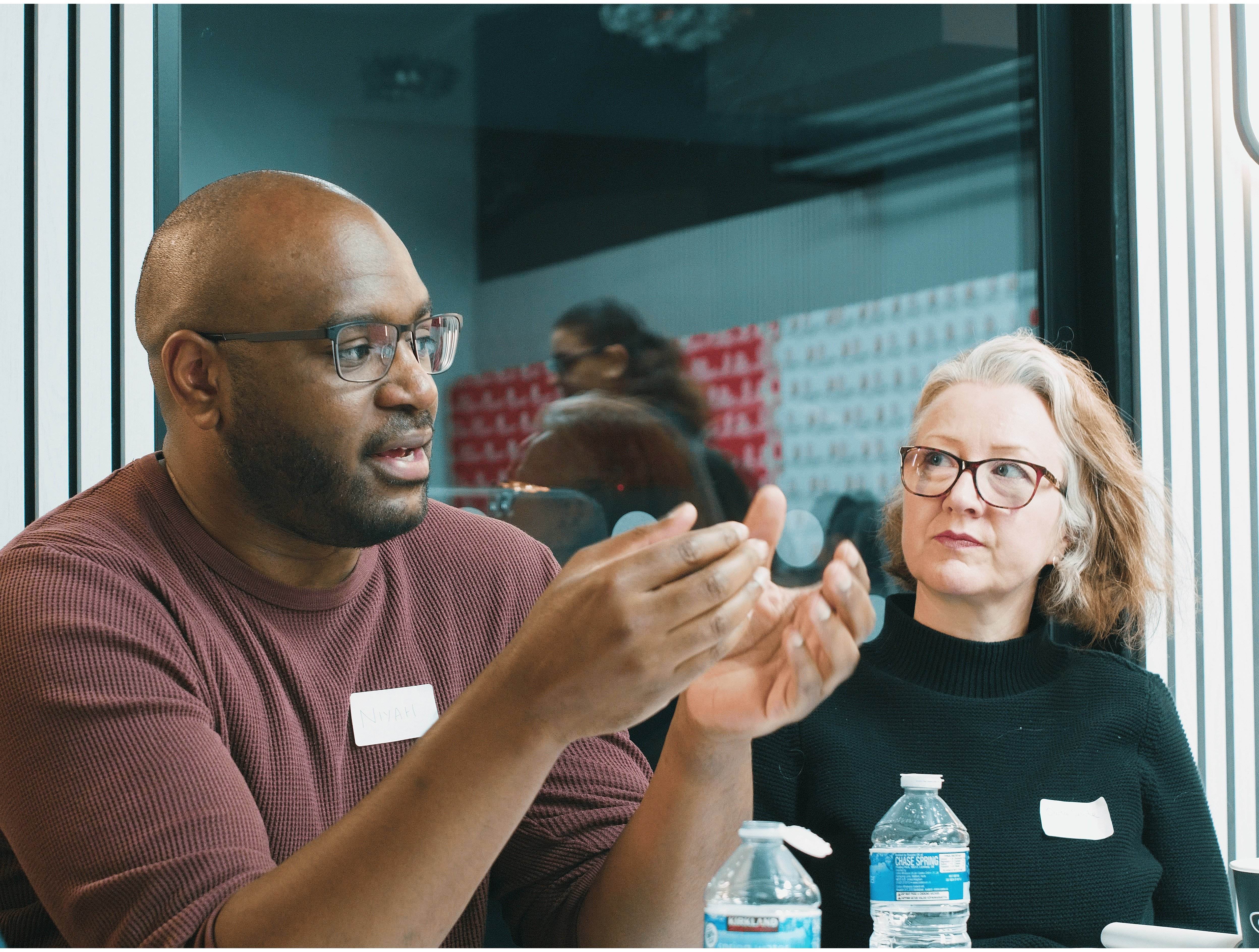 A man in glasses and a maroon sweater speaking while a woman with glasses and a black sweater listens attentively.